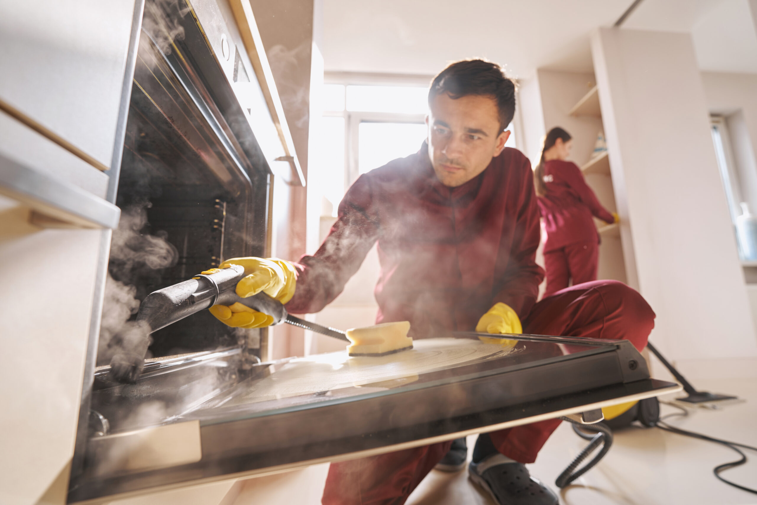 Cleaner using steam-cleaning device for disinfecting open oven door glass while his coworker dusting